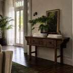 The Raffles Console in aged elm with rich grain, paired with a large plant, leaning framed artwork, and stacked books.