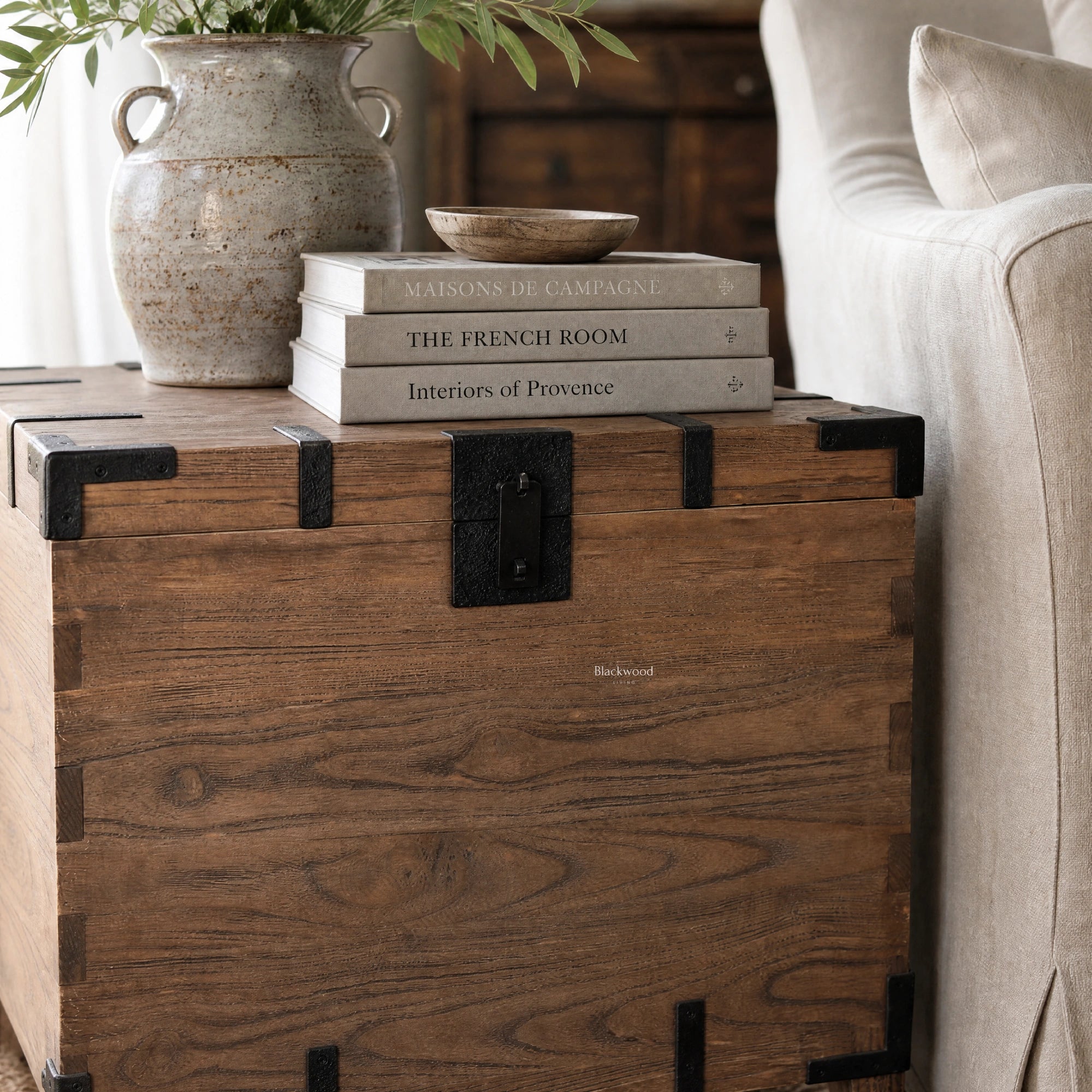 Avignon Trunk Table in reclaimed oak with exposed timber grain and forged metal details, beside books and a ceramic bowl.