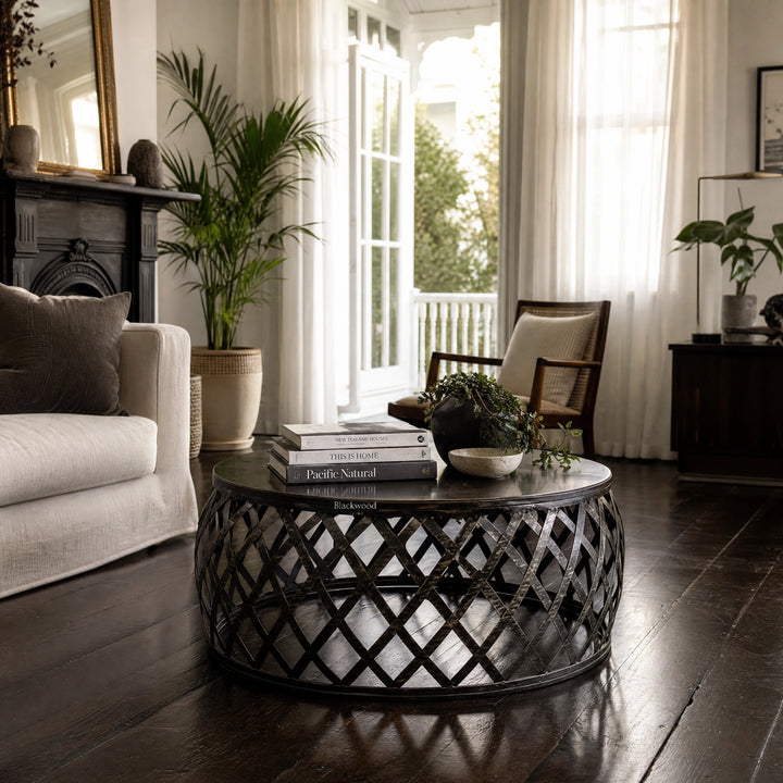 Mara Coffee Table with round iron lattice base in a sunlit living room, topped with books and plants.