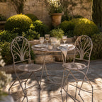 Provence Bistro Set in weathered antique grey with ornate scrollwork around a glass-top table in a sunlit courtyard.