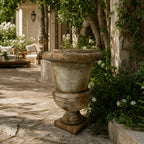 Provence Urn, antique marble-look magnesia composite, weathered finish, set in a sunlit stone courtyard with ivy.
