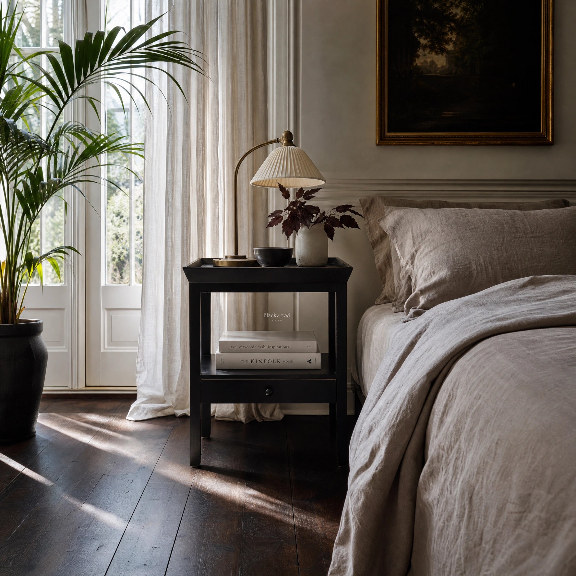 Colombo bedside table in black alder finish beside a bed with a pleated lamp, books, and greenery.