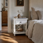 Bridgeport 1 Drawer Bedside in white with lower shelf, vase, bowls, and books beside a beige upholstered bed.