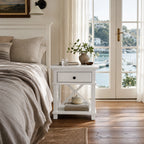 Bridgeport 1-Drawer Bedside in White beside a beige linen bed, with a potted plant and books on top.