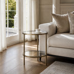Belgrave Side Table - Brass: brass-finished iron frame with two clear glass surfaces beside a beige sofa.