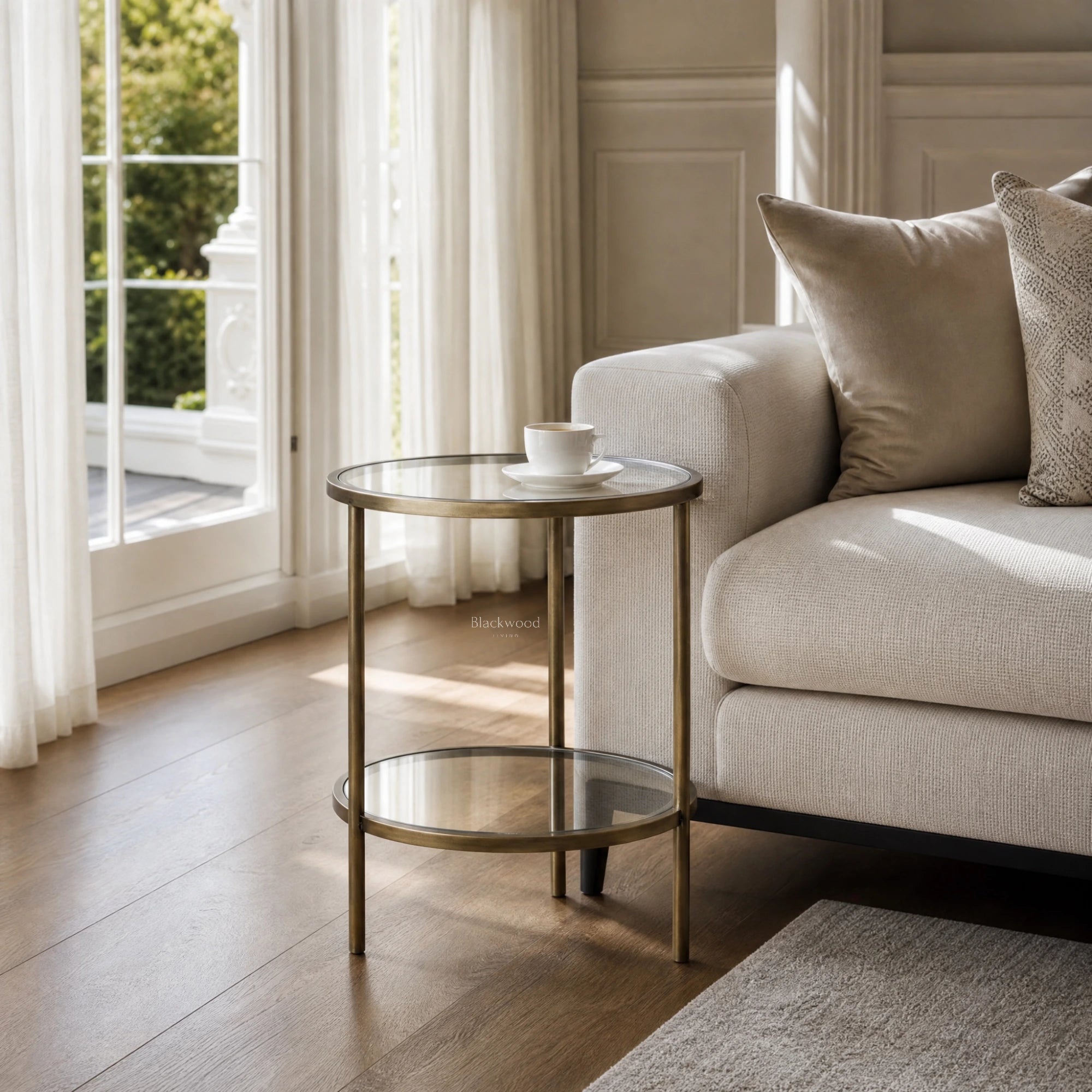 Belgrave Side Table - Brass: brass-finished iron frame with two clear glass surfaces beside a beige sofa.