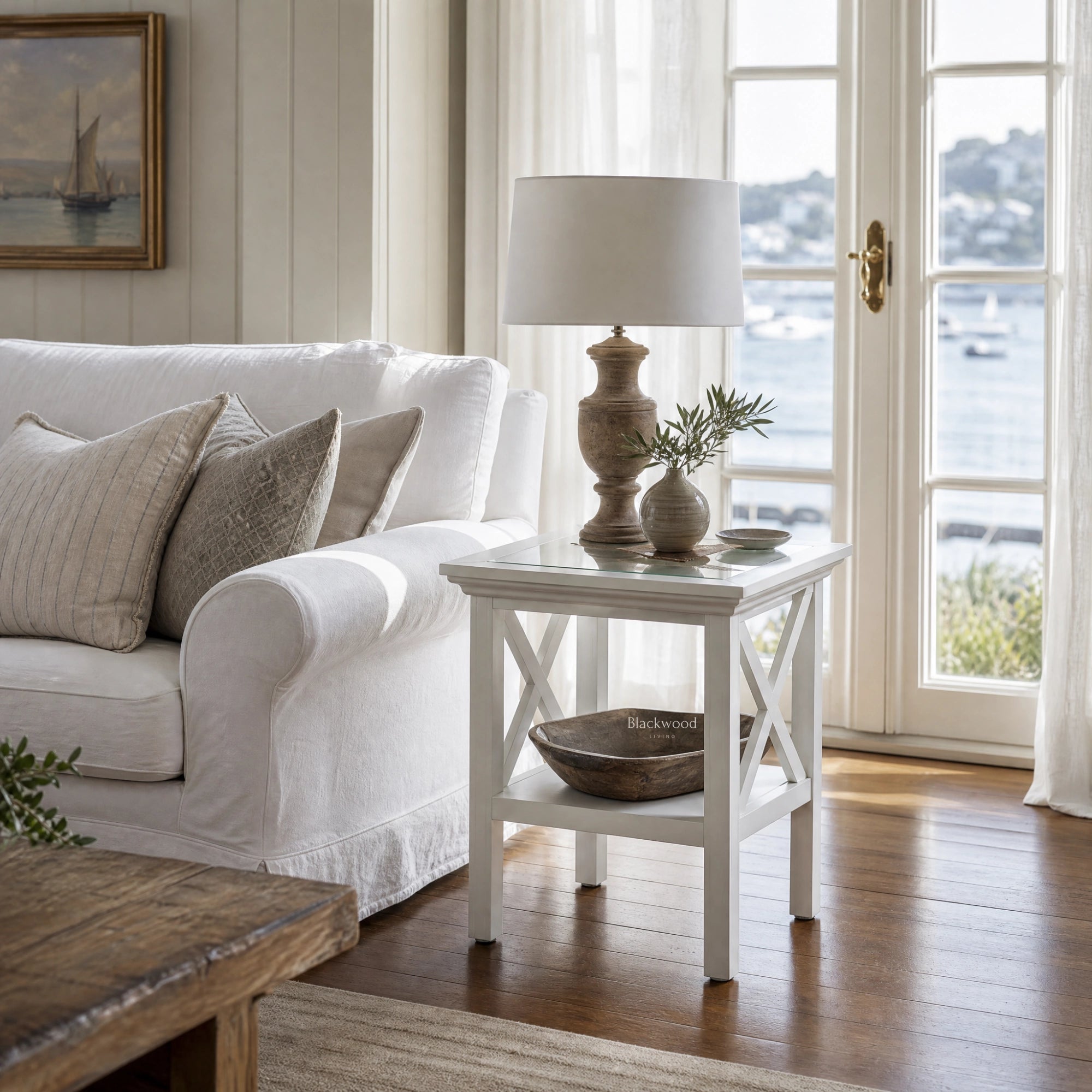 Newport Side Table in white with cross-X legs and lamp in a Hamptons-style living room by a water view.
