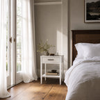 Rangoon White bedside table with two drawers and lower shelf beside a bed in a bright bedroom.
