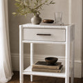 Rangoon Bedside Table - White with two drawers, topped by a vase, wooden bowl, books, and a glass.