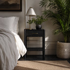 Rangoon Bedside Table in black with brass lamp and potted plant beside a white bed.