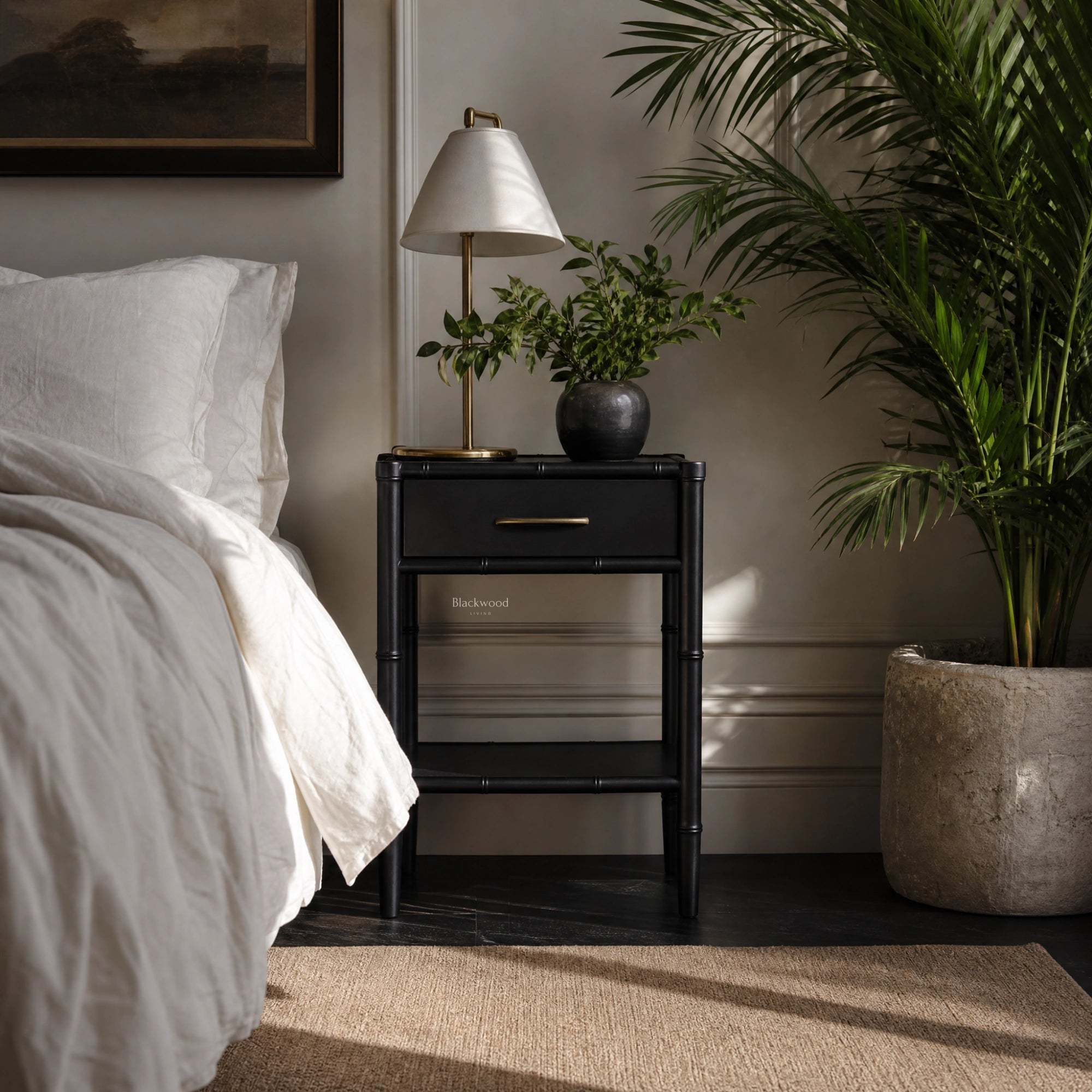 Rangoon Bedside Table in black with brass lamp and potted plant beside a white bed.