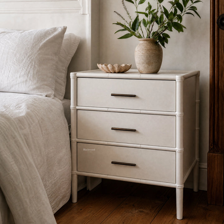 Rangoon 3-Drawer Bedside in white with bamboo detailing, topped by a speckled vase of greenery and a shell dish.