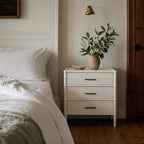 Rangoon 3 Drawer Bedside - White with brass handles, placed beside a bed and topped with a leafy plant in a vase.