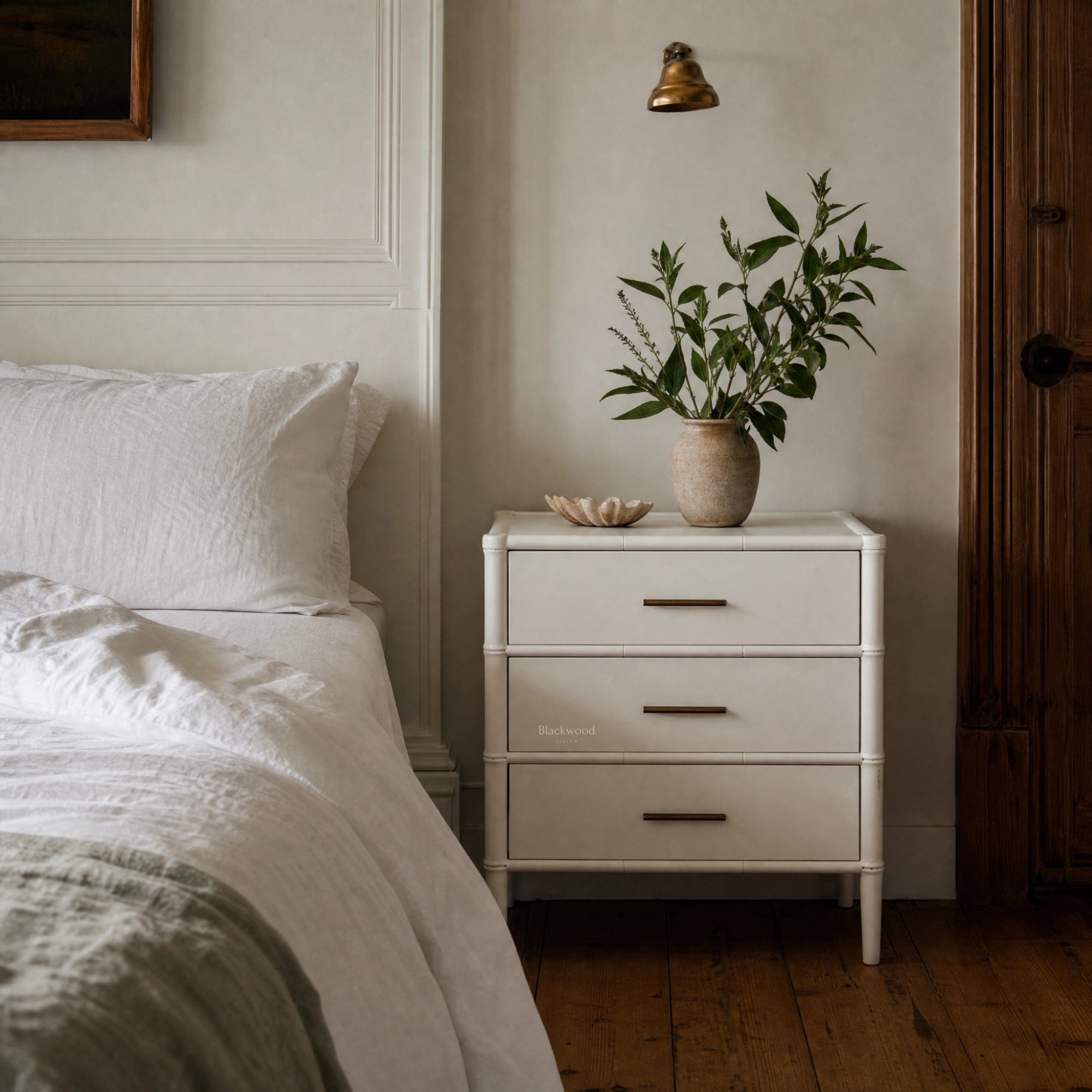 Rangoon 3 Drawer Bedside - White with brass handles, placed beside a bed and topped with a leafy plant in a vase.