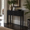 Black penang hall table with six drawers and warm metal handles in an entryway beside a vase and monstera plant.