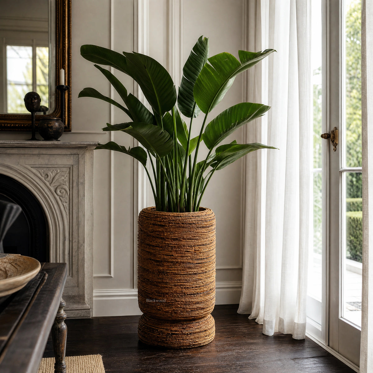 Large woven banana leaf planter holding a leafy green plant beside a marble fireplace and sheer curtains.