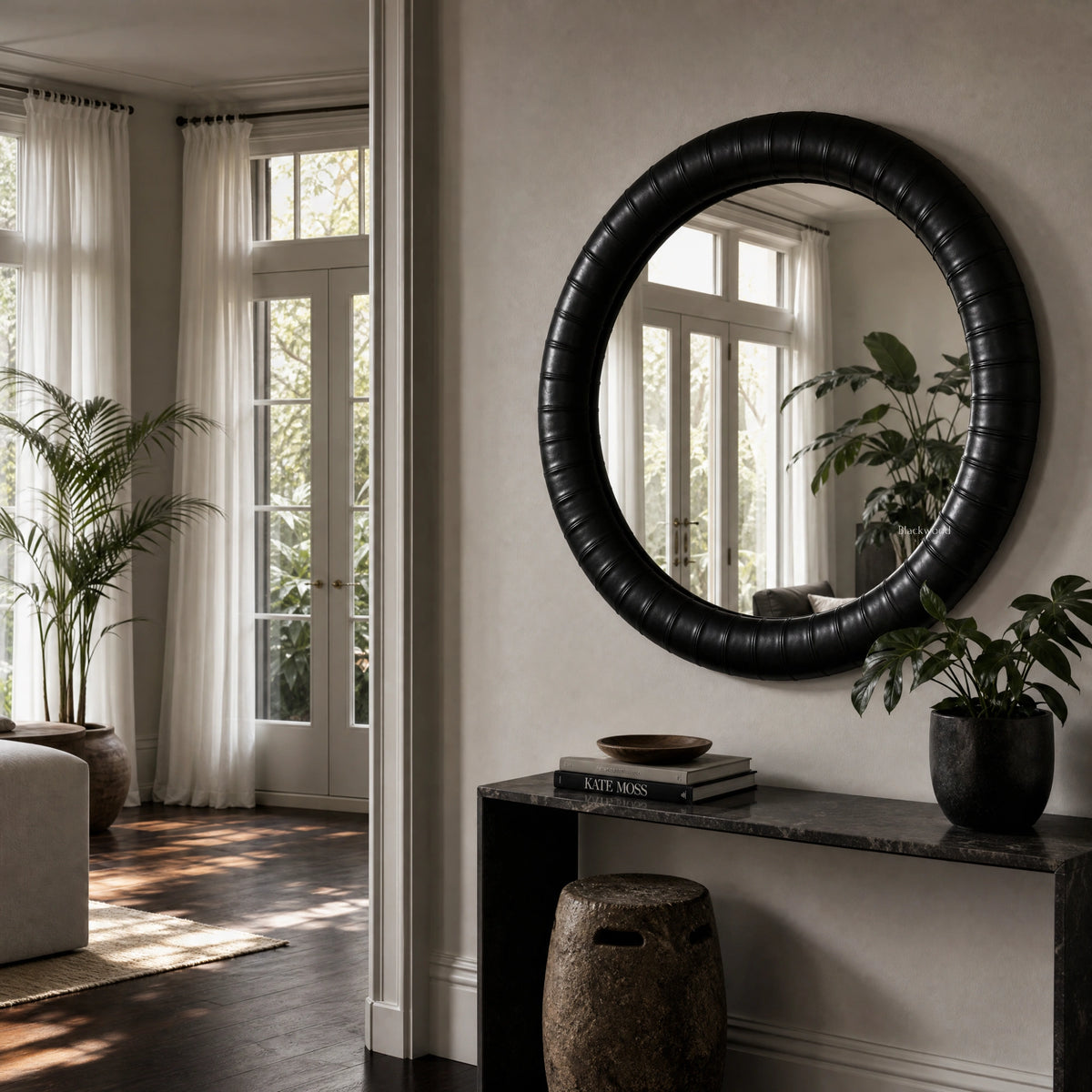Large round mirror with a thick, textured black leather frame featuring subtle panelled detailing, hanging above a dark console table.