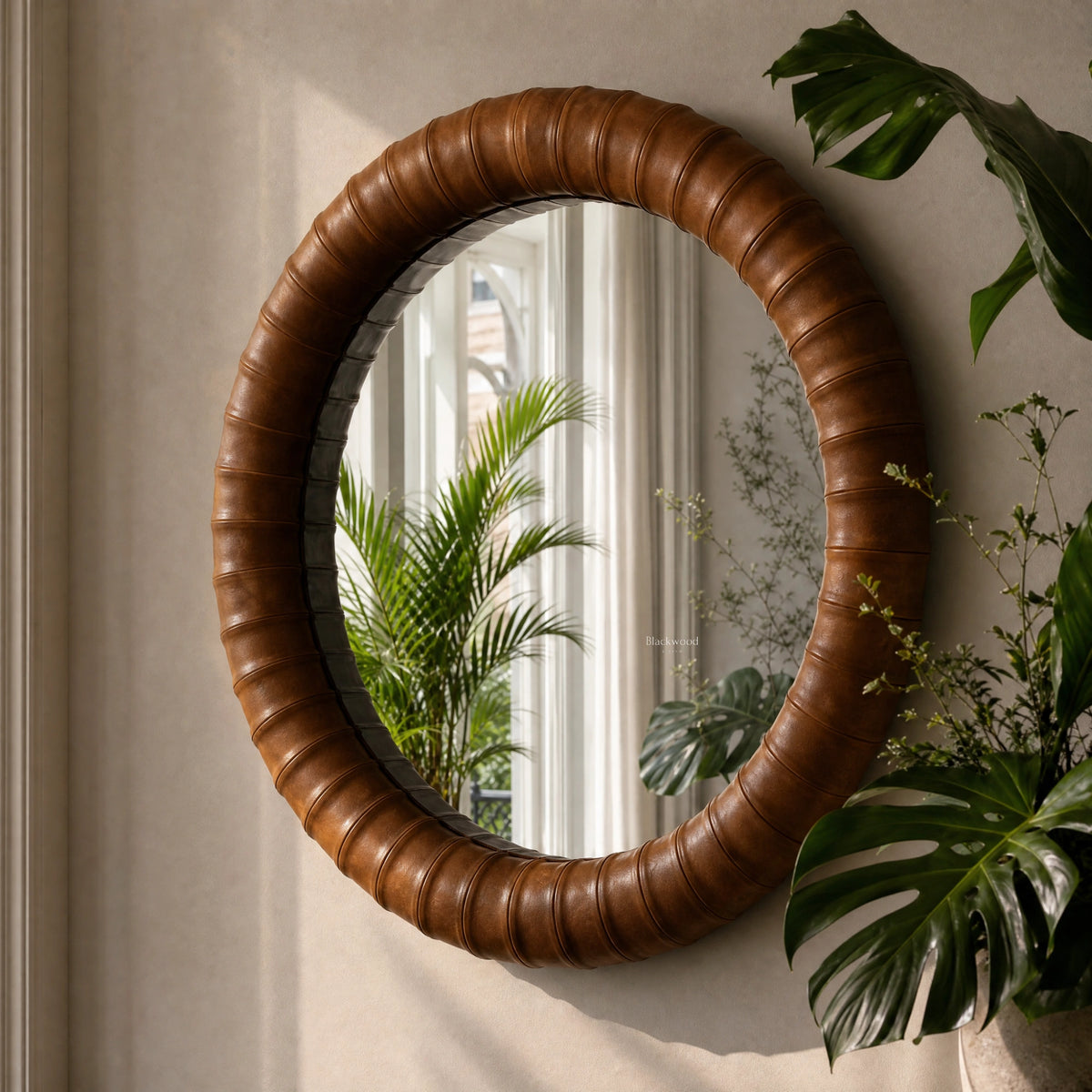 Round mirror with a thick, segmented cognac leather frame hanging on a wall beside tropical greenery.