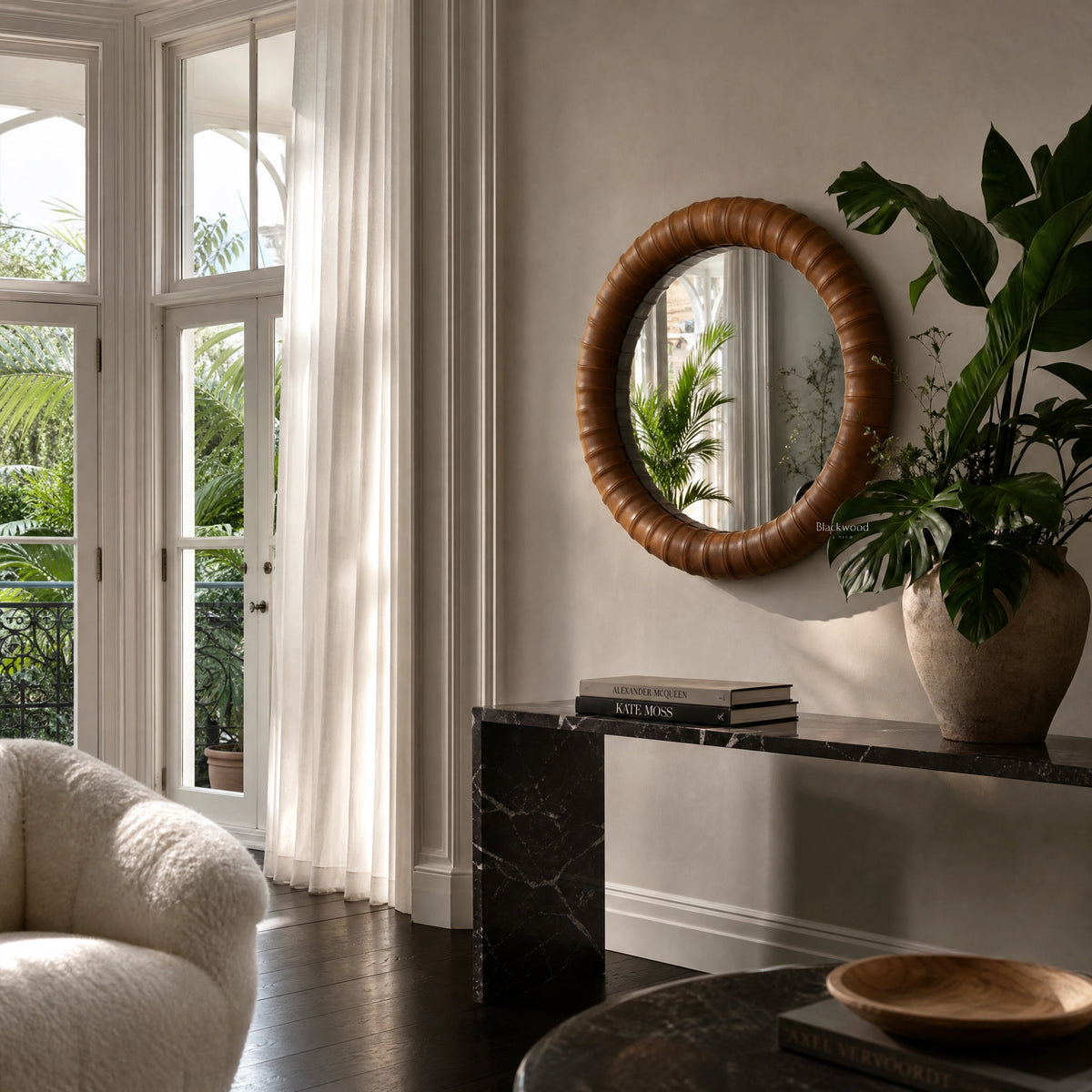 Round cognac leather mirror with ribbed frame hangs above a black marble console next to large green plants and sheer curtains.