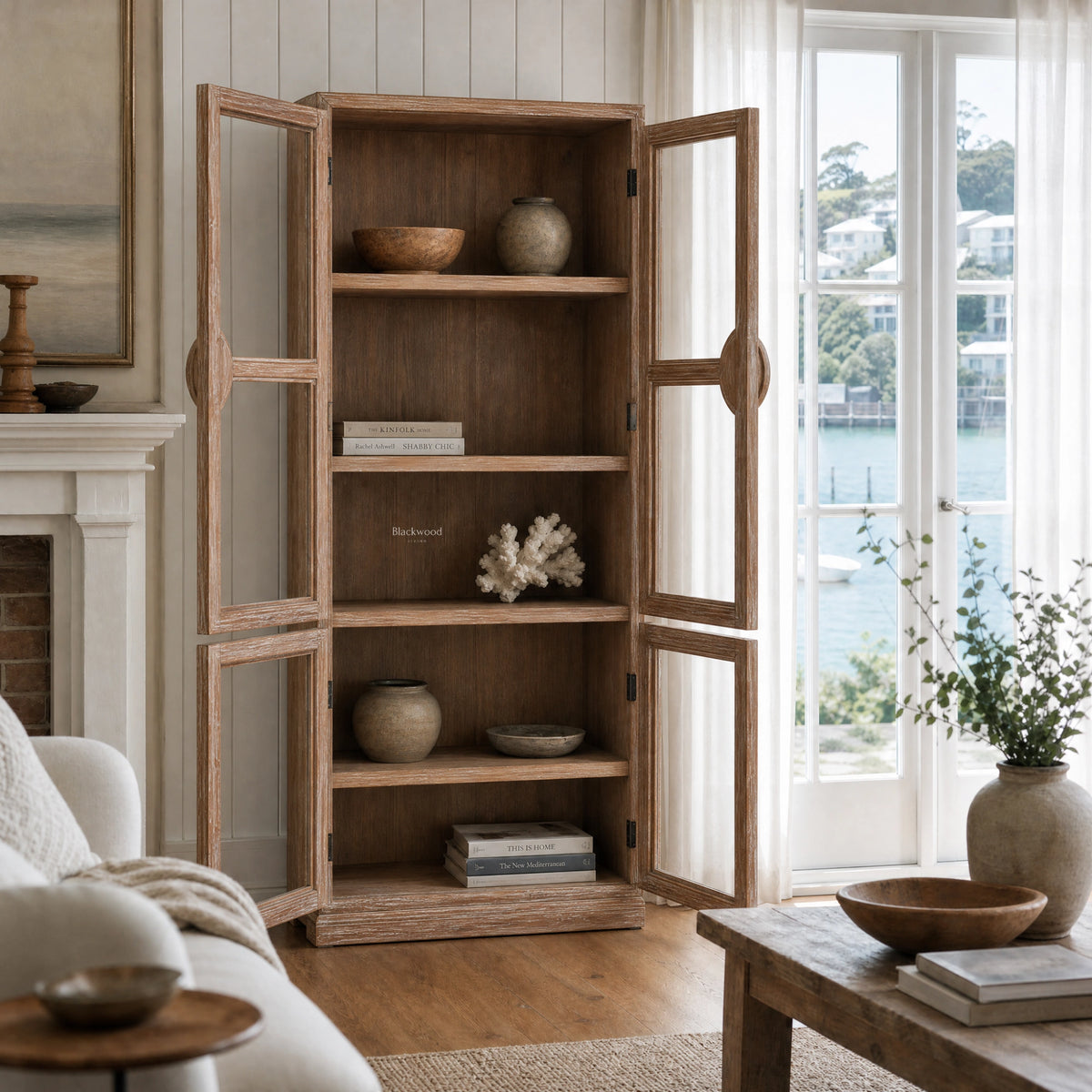 Tall elm Capstan Cabinet with open glazed doors, circular nautical handles, and shelves displaying books and pottery in a coastal living room.