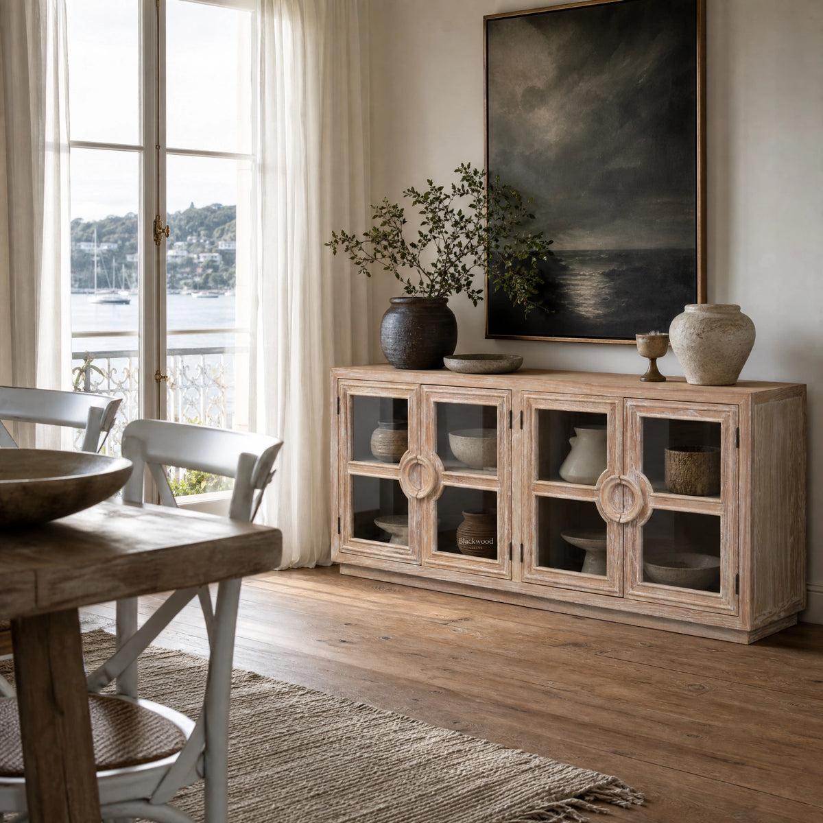 Rustic elm Capstan sideboard with glass doors and ring handles, styled with pottery and art near a coastal window view.