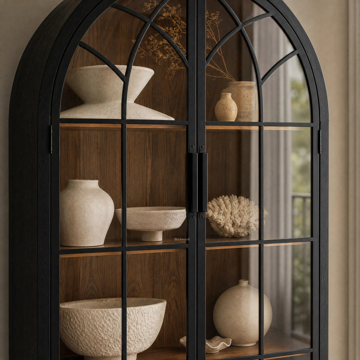 Tall arched cabinet with glazed doors displaying neutral ceramic vases and bowls on warm wood shelves within a dark frame.