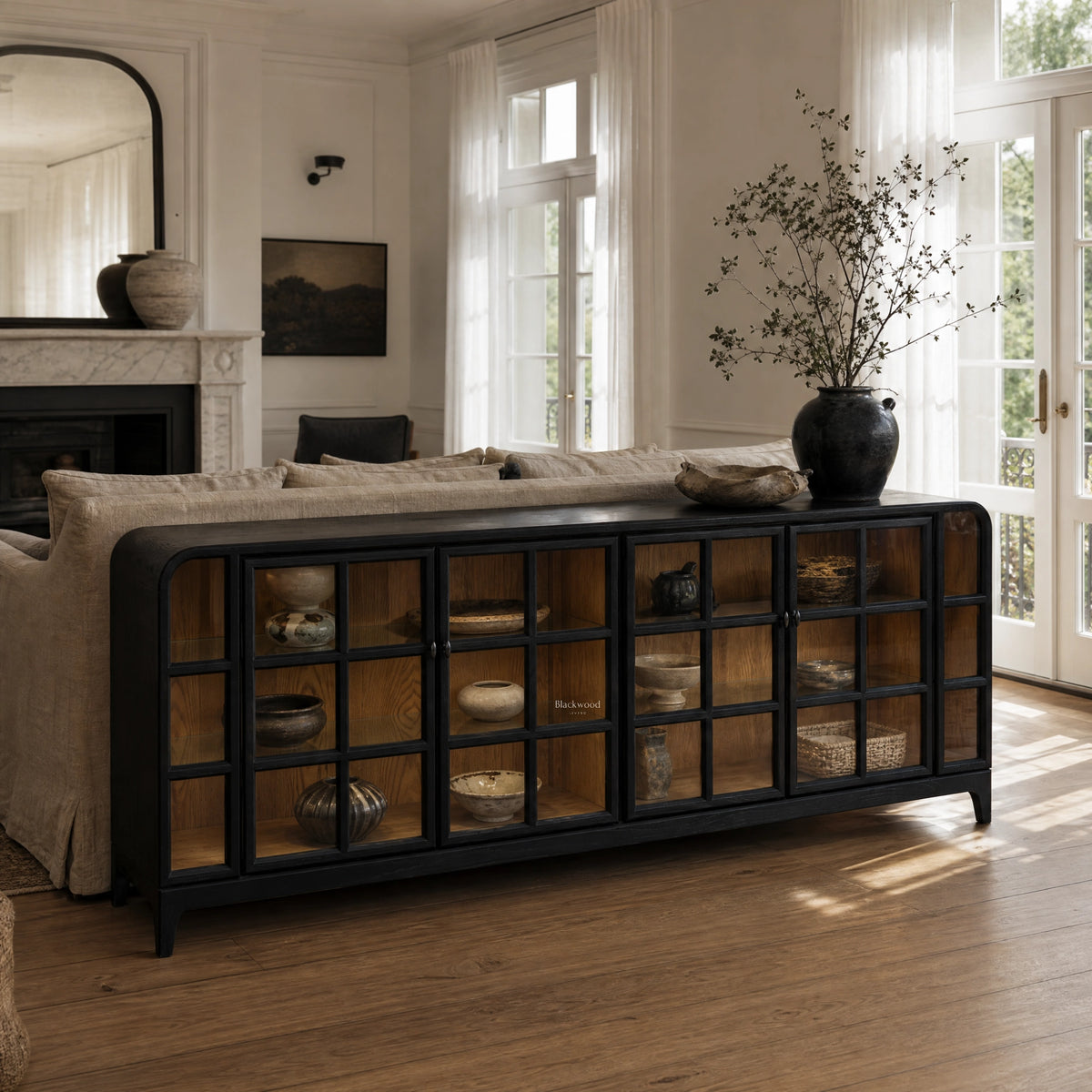 Dark glazed sideboard with grid glass doors and ash interior, anchoring a sunlit living room behind a linen sofa.