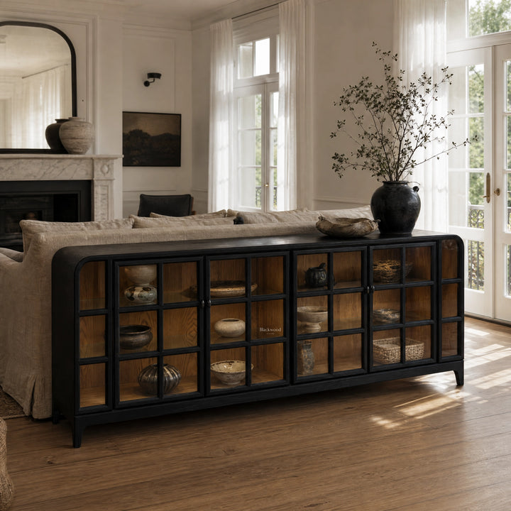 Dark glazed sideboard with grid glass doors and ash interior, anchoring a sunlit living room behind a linen sofa.