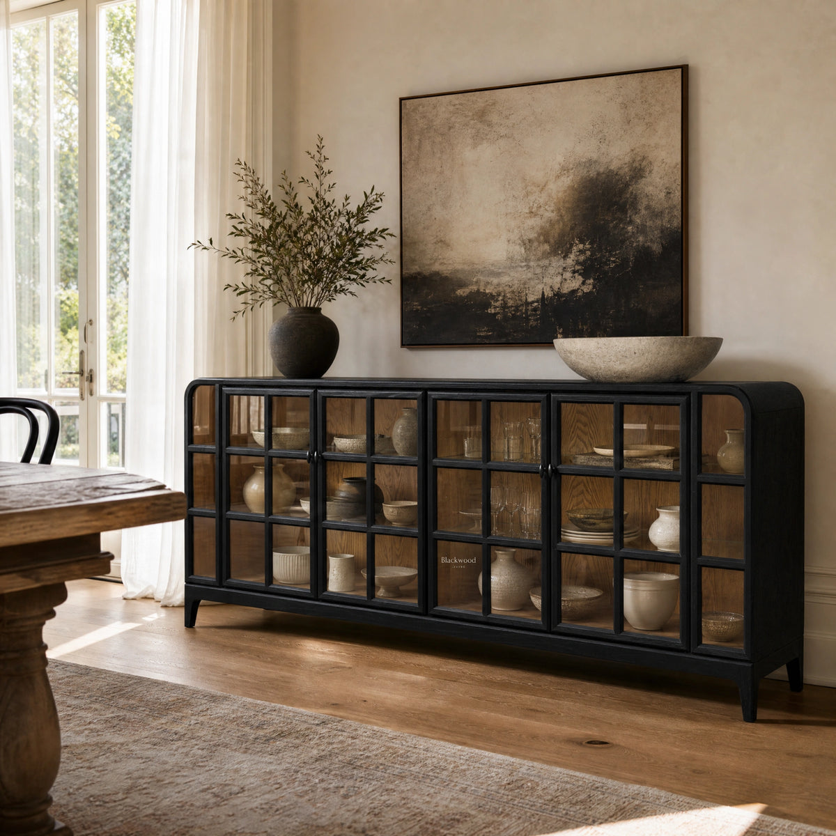 Dark ash sideboard with glazed grid doors displaying ceramic dishware in a sunlit dining room.