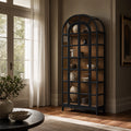 Tall arched display cabinet with black frame and glazed doors showcasing ceramic vases against warm wood interior in a sunlit room.