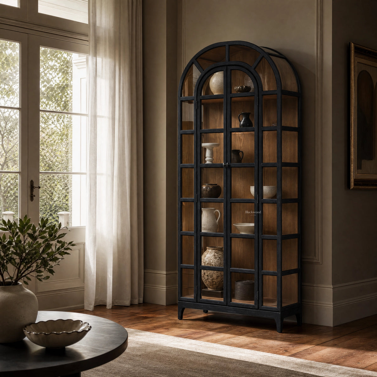 Tall arched display cabinet with black frame and glazed doors showcasing ceramic vases against warm wood interior in a sunlit room.