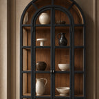 Arched glazed cabinet with dark frame and warm timber interior displaying ceramic vases and bowls. Heritage-style furniture.