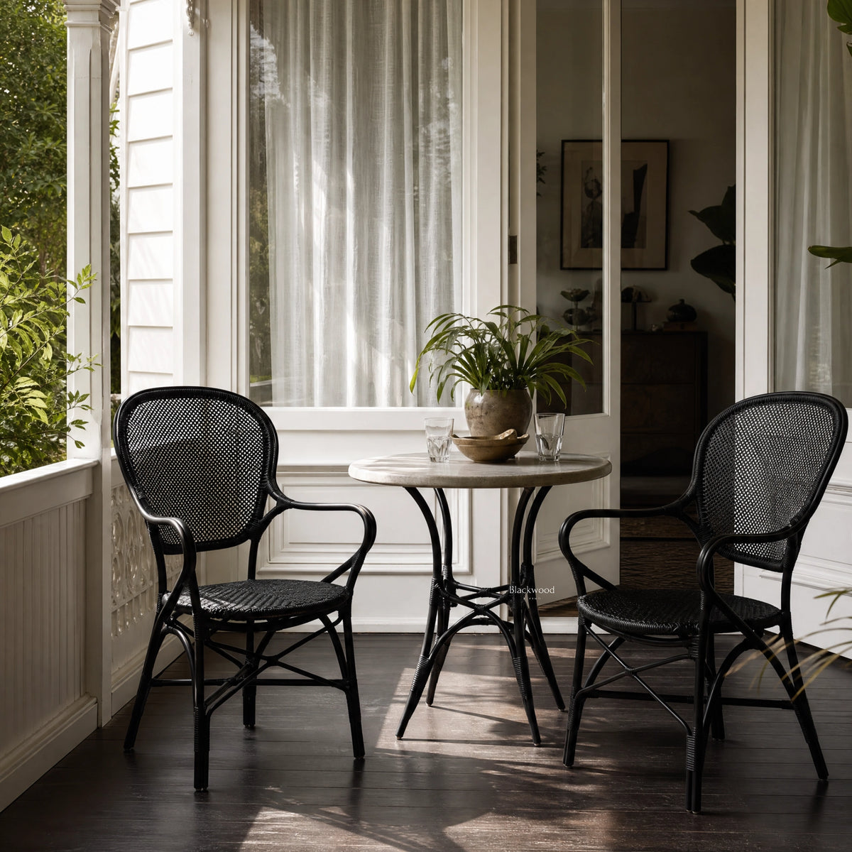 Two black Sika Rossini rattan armchairs flank a small round table on a sunlit porch with sheer white curtains.