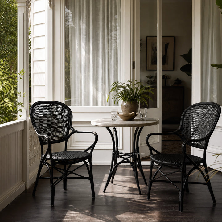 Two black Sika Rossini rattan armchairs flank a small round table on a sunlit porch with sheer white curtains.