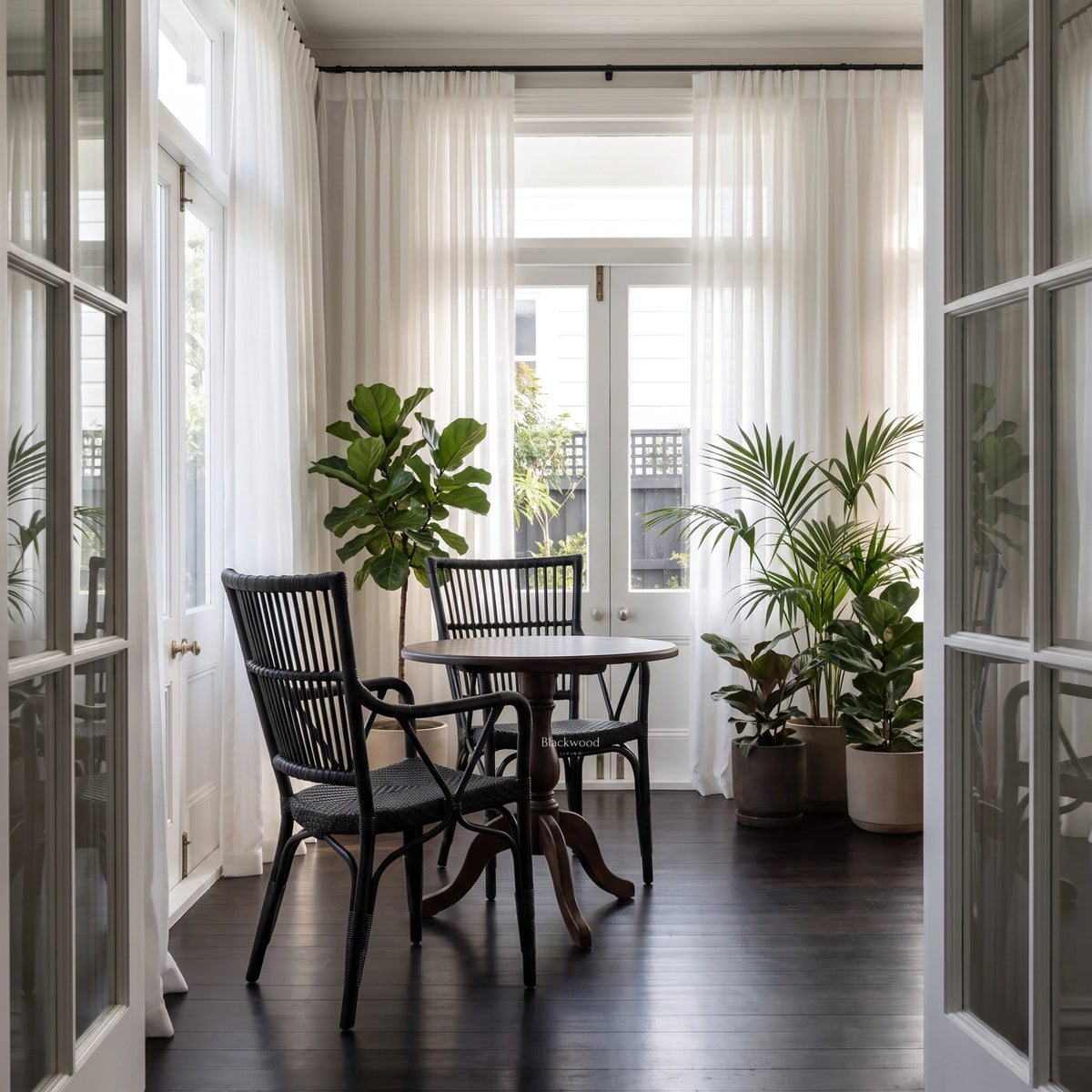 Black handmade rattan armchair with braided seat and vertical backrest beside a round table and potted plants in a sunlit room.