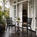 Two black Piano Rattan Armchairs with bar backrests and braided seats flank a small table on a porch with white columns.