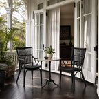 Two black Piano Rattan Armchairs with bar backrests and braided seats flank a small table on a porch with white columns.