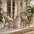 Two Sika Fleur rattan armchairs with lattice backs and linen cushions sit on a sunlit porch beside a stone side table.