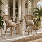 Two Sika Fleur rattan armchairs with lattice backs and linen cushions sit on a sunlit porch beside a stone side table.