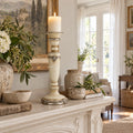 Tall cream candle holder with silver ornamental detailing and turned silhouette on a mantel beside rustic pottery and flowers.