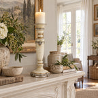 Tall cream candle holder with silver ornamental detailing and turned silhouette on a mantel beside rustic pottery and flowers.