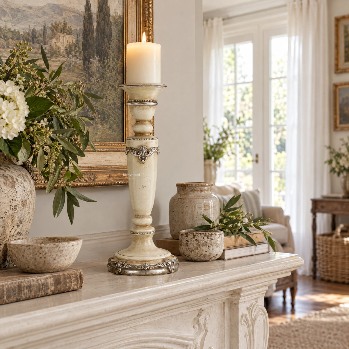 Tall cream candle holder with silver ornamental detailing and turned silhouette on a mantel beside rustic pottery and flowers.