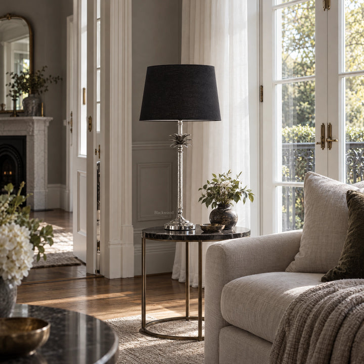 Tall polished metal palm table lamp with pineapple stem detail and deep black linen shade on a marble side table.