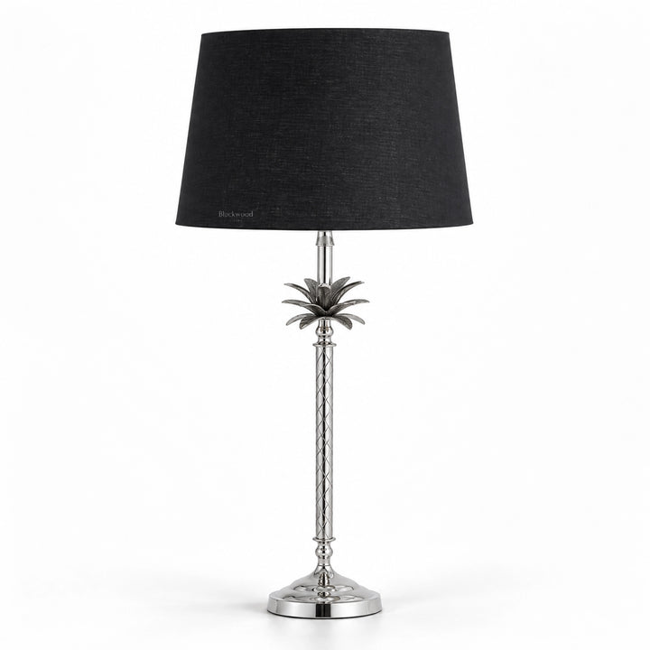 Tall polished metal table lamp with sculptural palm motif, diamond-patterned stem, and deep black linen shade.