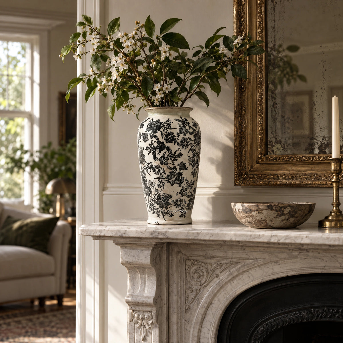 Tall ceramic vase with intricate black botanical detailing on warm ivory held by white blossoms on a marble mantelpiece.