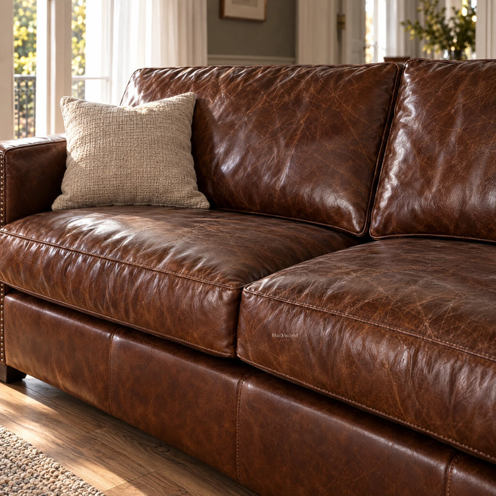 Brown leather three-seater sofa with vintage texture, plush cushions, and a beige throw pillow in a sunlit living room.