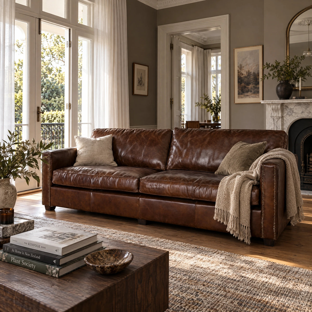 Brown leather 3 seat sofa with nailhead trim in a sunlit living room featuring a fireplace and wooden coffee table.