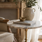 Marcel Side Table with veined marble top on an antique gold pedestal, shown beside a ceramic vase with greenery.