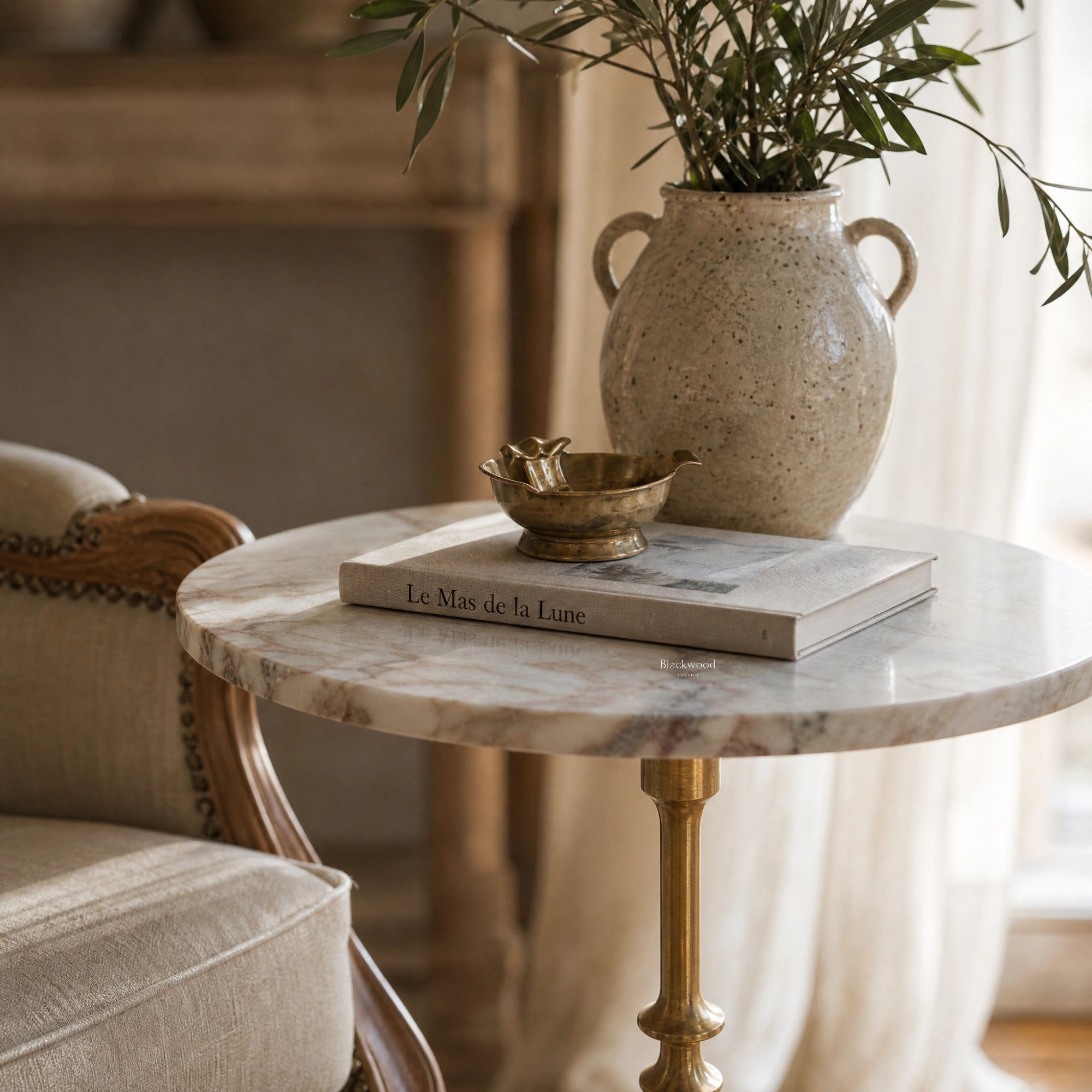 Marcel Side Table with veined marble top on an antique gold pedestal, shown beside a ceramic vase with greenery.