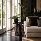Banyan Side Table in cacao-stained solid oak with a slatted pedestal beside a sofa in a sunlit living room.